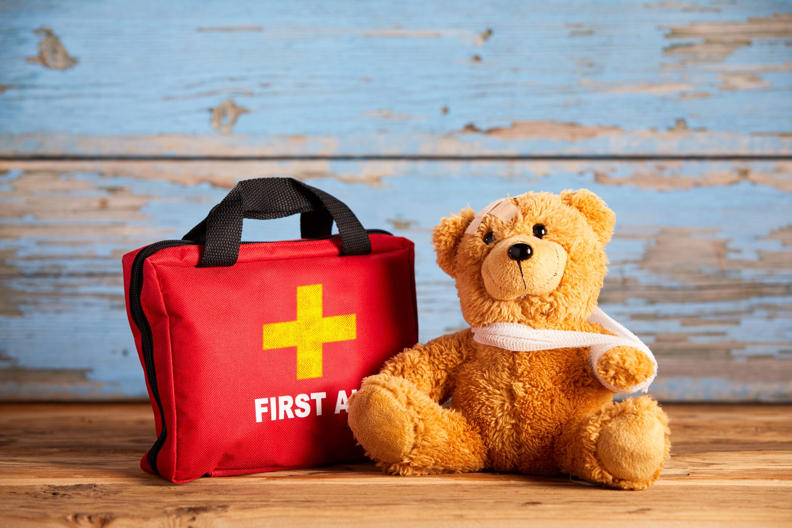 Little teddy bear with an injured arm in a sling sitting alongside a red First Aid bag on rustic wood in a concept of paediatric healthcare and emergency triage